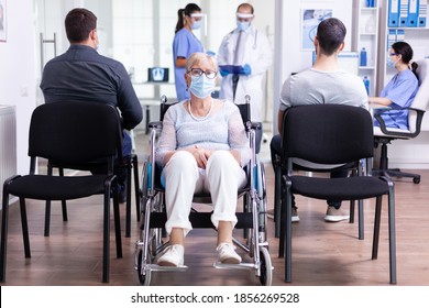 Worried Disabled Senior Woman Sitting In Wheelchair In Hospital Waiting Area For Doctor Examination. Old Woman Wearing Face Mask Against Coronavirus Infection.