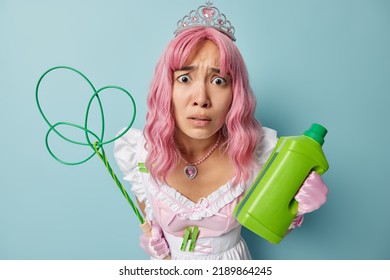 Worried Displeased Housewife With Pink Hair Holds Green Bottle Of Detergent And Carpetbeater Busy Doing Cleaning At Home Wears White Dress And Crown Isolated Over Blue Background. Housework Concept