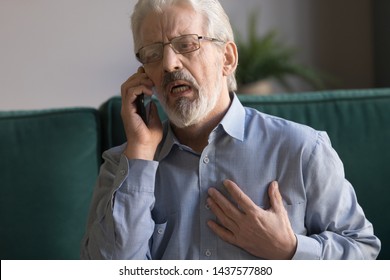 Worried elder senior man talking on phone calling for help 911 emergency ambulance, upset old middle aged grandfather touching chest feel sudden pain heartburn having heart attack sit on sofa at home