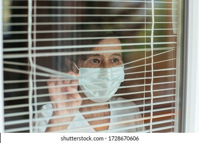 Worried elderly woman wearing face mask looking through window blinds during self-quarantine. Senior female wearing surgical mask looking outside the window at home. - Powered by Shutterstock