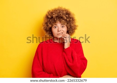 Worried embarrassed woman with Oops expression looks scared and guilty bites lips keeps fingers on chin looks nervously at camera dressed in red pullover isolated against yellow background