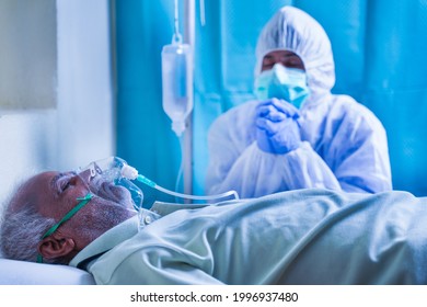 Worried Family Member Or Doctor In PPE Or Protective Suit Praying For Coronavirus Or Covid-19 Infected Patient For Recovery At Hospital While Patient Breathing On Ventilator Oxygen Mask