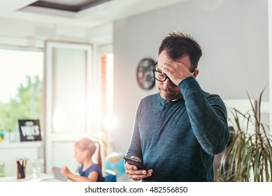 Worried father looking at smart phone and holding hand on forehead, in background his daughter doing homework - Powered by Shutterstock