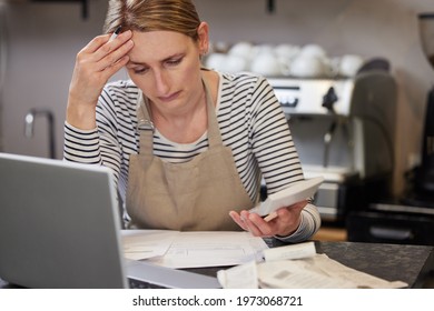 Worried Female Owner Of Coffee Shop In Financial Difficulty Looking Through Bills Using Laptop And Calculator