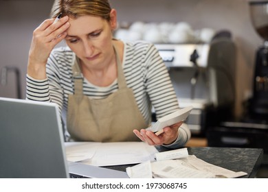 Worried Female Owner Of Coffee Shop In Financial Difficulty Looking Through Bills Using Laptop And Calculator