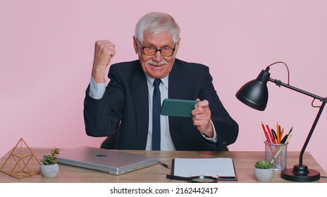 Worried Funny Senior Businessman Sits At Workplace Avoid Working And Enthusiastically Playing Racing Or Shooter Video Games At Office Desk Table. Elderly Man Isolated Indoor On Pink Studio Background