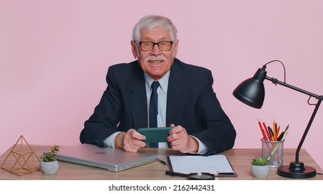 Worried Funny Senior Businessman Sits At Workplace Avoid Working And Enthusiastically Playing Racing Or Shooter Video Games At Office Desk Table. Elderly Man Isolated Indoor On Pink Studio Background