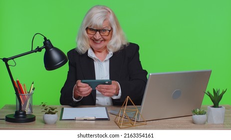 Worried Funny Senior Businesswoman Sits At Workplace Avoid Working And Enthusiastically Playing Racing Or Shooter Video Games At Office Desk Table. Elderly Woman On Green Chroma Key Studio Background