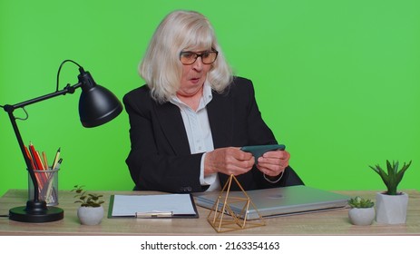 Worried Funny Senior Businesswoman Sits At Workplace Avoid Working And Enthusiastically Playing Racing Or Shooter Video Games At Office Desk Table. Elderly Woman On Green Chroma Key Studio Background