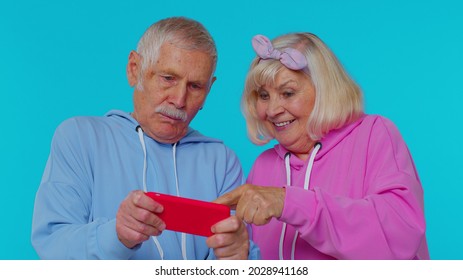 Worried Funny Senior Old Gray-haired Grandfather Grandmother Enthusiastically Playing Racing Video Games On Mobile Phone Isolated On Blue Studio Background. Mature Man Woman Grandparents Pensioners