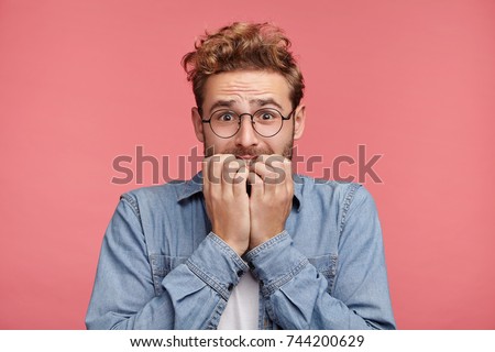 Worried hipster guy bites nails, looks nervous before passing exam or important event in his life. Embarrassed fashionable young man being afraid of difficulties, stands against pink background