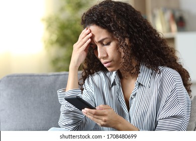 Worried latin woman reading on smart phone bad news sitting on a couch at home