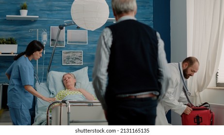 Worried Man Calling For Medical Assistance While Woman Breathing Heavy In Hospital Bed. Doctor And Nurse Rushing To Help Retired Patient With Disease For Healthcare In Nursing Home.