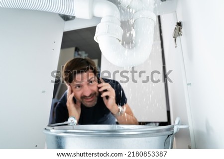 Worried Man Calling Plumber While Watching Water Leaking From Sink