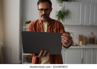 Worried Man In Eyeglasses Looking At Laptop In Blurred Kitchen