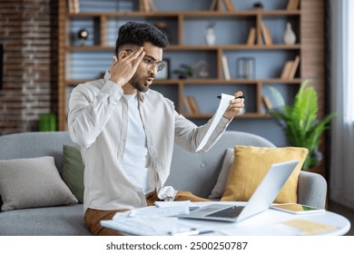 Worried man holding receipt sitting on couch with laptop, documents, and phone. Man appears stressed checking expenses or bills in home environment. - Powered by Shutterstock