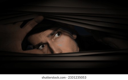 Worried man looking through window blinds into darkness. Paranoia concept