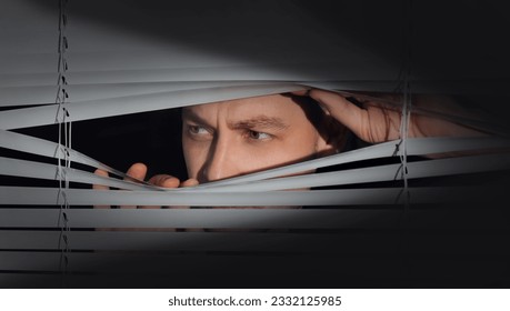 Worried man looking through window blinds into darkness. Paranoia concept
