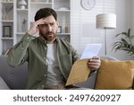 Worried man reading letter on couch at home, holding envelope, showing signs of stress and anxiety. Concerned expression indicates financial issues or bad news.