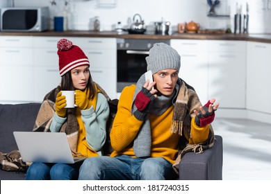 Worried Man Talking On Smartphone Near Cold Woman Wearing Warm Hat Nad Gloves While Sitting With Laptop In Cold Kitchen