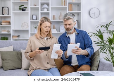 Worried mature couple reviewing bills together at home, calculating finances with a smartphone and notepad. Concept of financial planning and family budgeting. - Powered by Shutterstock