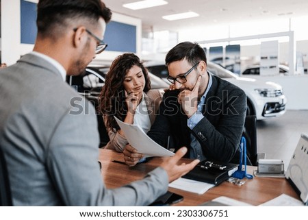 Worried middle age man with his wife buying car in auto salon or showroom. He is hipped while woman asks him to still buy a car.