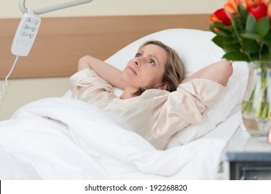 Worried middle-aged woman lying thinking in a hospital bed with her hands clasped behind her head as she stares at the ceiling with an intense expression - Powered by Shutterstock