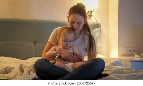 Worried Mother Checking Body Temperature Of Her Sick Baby Son Awake At Night.