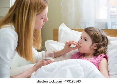 Worried Mother Giving Glass Of Water To Her Ill Kid. Sick Child With High Fever Laying In Bed And Taking A Medicine. Hand On Forehead. 