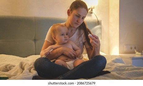 Worried Mother Looking At Thermometer After Measuring Temperature Of Her Sick Baby Son At Night.