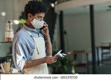 Worried New Business Owner Calculating Finances. Young African American Male In Apron And Protective Mask Looks At Bills And Speaks On Phone In Modern Cafe Or Restaurant Interior, Copy Space