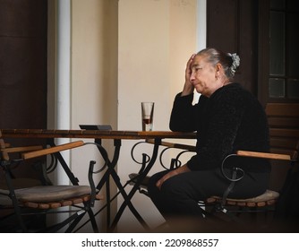 Worried Old Woman Sits At A Bar Table. She Rests Her Head On Her Palm. A Glass Of Drink On The Table. France, Paris.September, 29, 2022