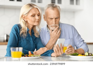 Worried Older Spouses Looking At Smartphone And Having Breakfast In Kitchen, Stressed Senior Couple Reading News Online Or Checking Incoming Message, Sitting At Table At Home, Closeup - Powered by Shutterstock