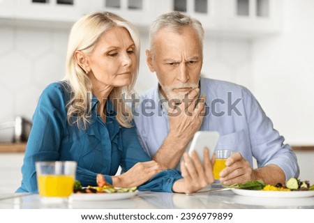 Worried Older Spouses Looking At Smartphone And Having Breakfast In Kitchen, Stressed Senior Couple Reading News Online Or Checking Incoming Message, Sitting At Table At Home, Closeup