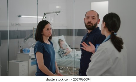 Worried Parents Discussing Sickness Recovery Treatment With Pediatrician Doctor In Hospital Ward. Hospitalized Girl With Nasal Oxygen Tube Sleeping After Disease Surgery During Medical Examination