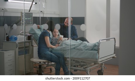 Worried Parents Sitting With Sick Girl Daughter Waiting For Sickness Expertise During Medical Examination In Hospital Ward. Child Wearing Oxygen Nasal Tube Holding Teedy Bear While Resting In Bed