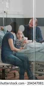 Worried Parents Sitting With Sick Girl Daughter Waiting For Sickness Expertise During Medical Examination In Hospital Ward. Child Wearing Oxygen Nasal Tube Holding Teedy Bear While Resting In Bed