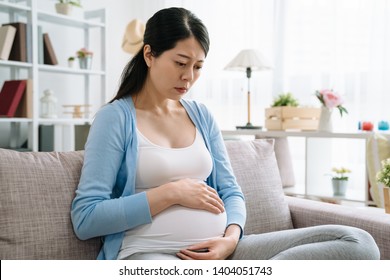 Worried Pregnant Woman Sitting On Sofa At Home Frowning Face Considering Problem. Asian Girl With Baby In Belly Sitting On Couch Upset Sadness Resting In Living Room Indoors. Stressed Motherhood.