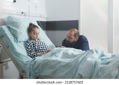 Worried sad uneasy father watching sick little daughter sleeping in hospital bed while in pediatric clinic patient room. Under treatment ill child resting while attentive parent taking care of her. - Powered by Shutterstock