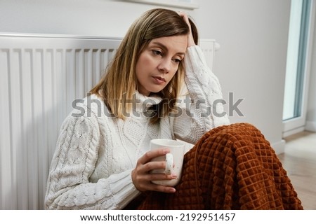 Worried sad woman sits under blanket near heating radiator with cup of tea, Rising costs in private households for gas bill due to inflation and war, Energy crisis