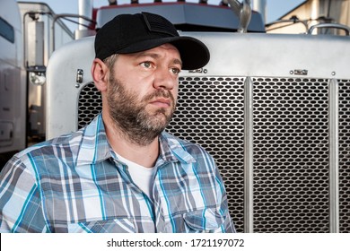 Worried Semi Truck Driver Wearing Plaid Shirt And Black Baseball Hat Sits In Front Of Big Rig. American Trucker With A Thoughtful Expression Sits In Front Of Big Rig. Concept Of Trucking Business.