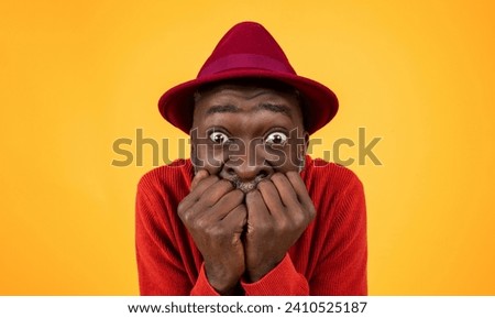 Worried senior Black man in a red sweater and hat, biting his nails with wide eyes full of anxiety against a plain yellow background, showing a humorous expression of suspense