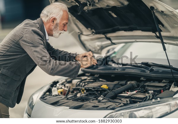 Worried senior business\
man taking photos with smartphone while trying to see what\'s wrong\
with his car.