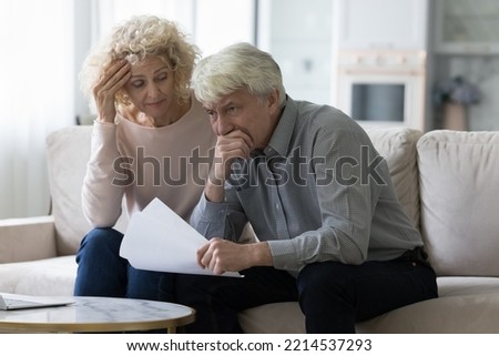 Worried senior wife and husband sit on sofa hold papers with unpleasant news, got notification from bank having financial problems, looking concerned. High taxes, eviction, late payments, subpoenas