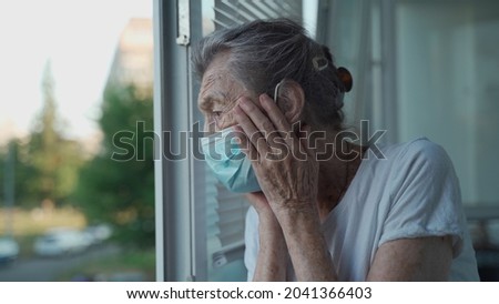 Worried senior woman in a protective medical mask sadly looks out the window with her head clasped in her hands in a nursing home. Depressed lady at home during the covid-19 pandemic.