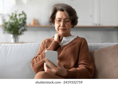 Worried senior woman sitting on sofa at home and looking at phone screen, reading or sending messages, video calling to family or watching news - Powered by Shutterstock