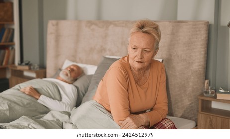 Worried senior woman sitting on bed with her husband sleeping in background - Powered by Shutterstock