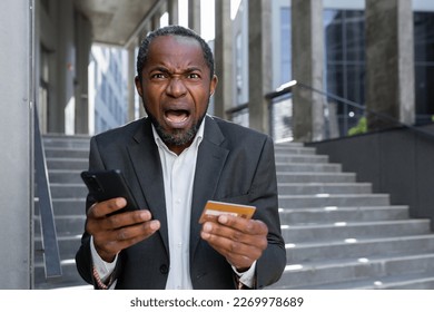 Worried shocked senior african american businessman in suit standing outside near office center. Holds a blocked credit card and phone. Bankruptcy, fraud. Screams angrily at the camera. - Powered by Shutterstock