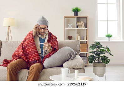 Worried sick man with seasonal cold or flu fever symptoms takes his temperature. Sad unhappy man in woolen hat, scarf and warm plaid sitting on couch at home and looking at thermometer in his hand - Powered by Shutterstock