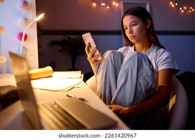 Worried Teenage Girl Sitting At Desk In Bedroom At Home Looking At Mobile Phone At Night